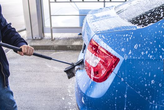 Manual car wash with pressurized water in car wash outside.Using a brush to wash a car on a car washing facility on sunny summer day.Blue car manual wash of with pressurized water in car wash outside.