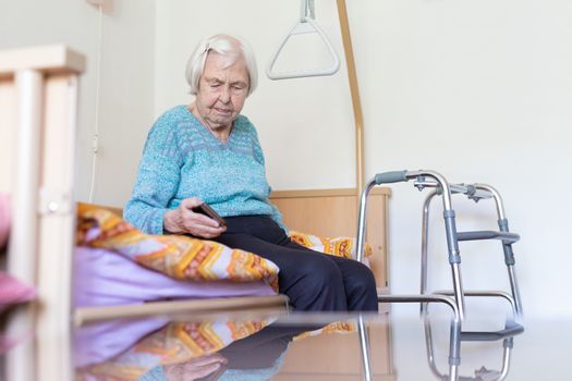 Elderly 96 years old woman reading phone message while sitting on medical bed supporting her by holder. Senior woman using wireless internet connection on smart phone.