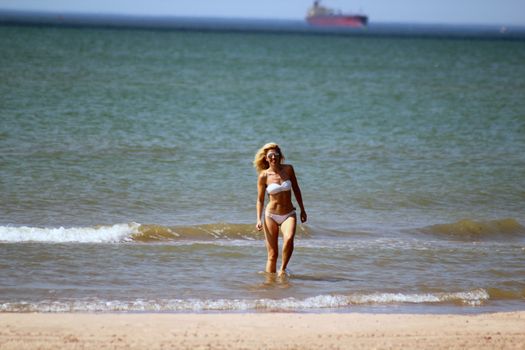 tanned blonde in white bikini and sunglasses comes out of the water on the sand of the beach after swimming in the Baltic Sea. Behind against the background a ship in the sea is visible in the distance. Sunny day.