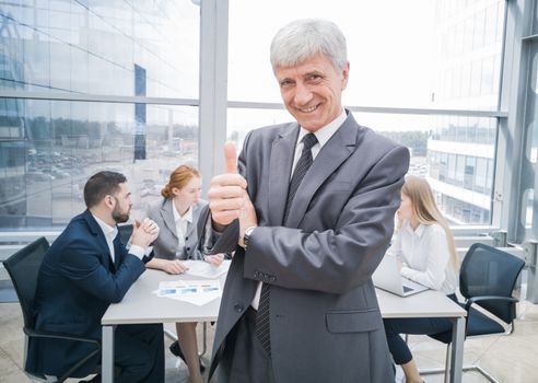 Confident mature businessman showing thumb up and smiling , team of his colleagues sitting at the table in the background