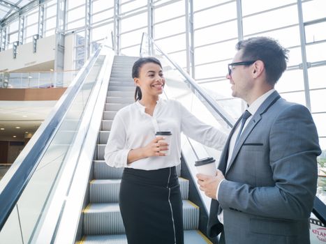 Businessman and businesswoman standing together on escalator and talk about business holding coffee in hand