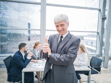 Confident mature businessman showing thumb up and smiling , team of his colleagues sitting at the table in the background