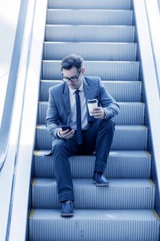 Businessman sitting on escalator surfing the net and holding coffee in hand