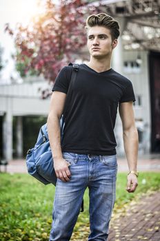 Attractive young man wearing black T-shirt and jeans with backpack standing outside