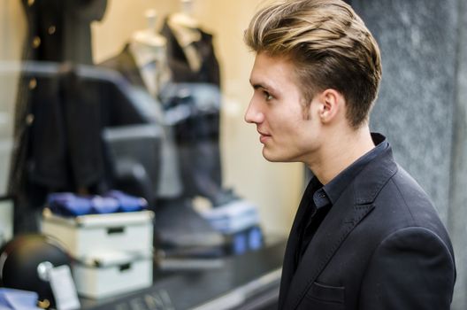 Handsome Young Man in Black Elegant Suit Looking at Displayed Fashion Items in Glass Window Boutique at the Street Side.
