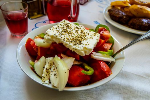 Greek salad of fresh cucumber, tomato, sweet pepper, lettuce, red onion, feta cheese and olives with olive oil in a white plate on a white table. View from above.