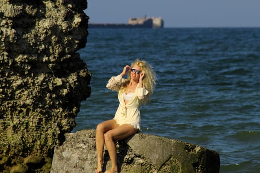 tanned blonde in white dress and sunglasses sits on a stone on the shore of the Baltic Sea. Sunny day