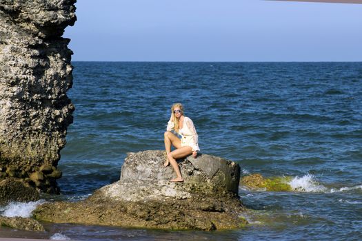 tanned blonde in white dress and sunglasses sits on a stone on the shore of the Baltic Sea. Sunny day