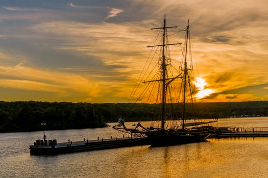 A two-mast brig stands moored in the harbor in the rays of the setting sun, painted the sky and everything around in yellow