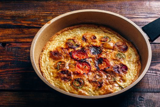 Cooked frittata with chorizo, tomatoes and chili peppers in a frying pan over dark wooden background. High angle view.