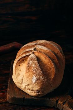 Homemade rye loaf on cutting board with knife over dark wooden background.