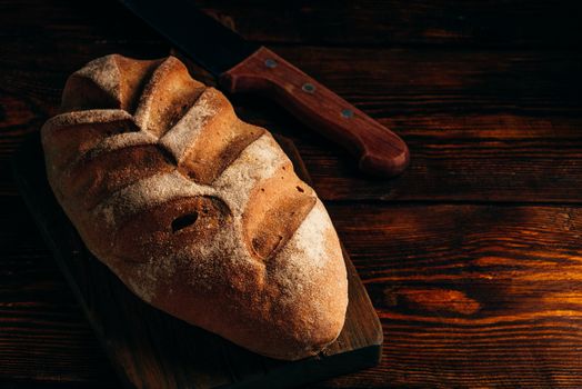 Homemade loaf on cutting board with knife over dark wooden background.