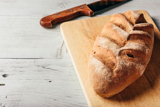 Homemade rye loaf on cutting board with knife over light wooden background.