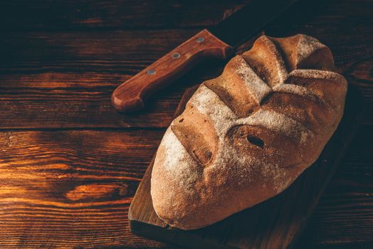 Homemade rye loaf on cutting board with knife over dark wooden background.