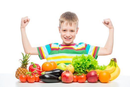 Boy athlete shows biceps at the table with a pile of fresh vegetables and fruits isolated in studio