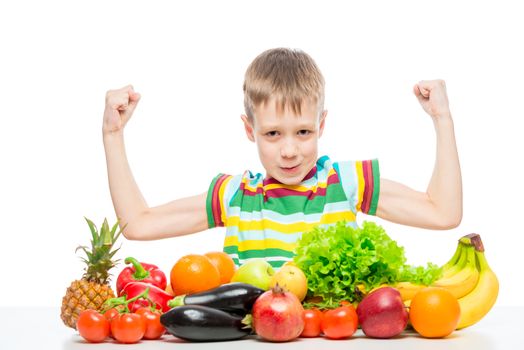 small strongman shows biceps at the table with a pile of fresh vegetables and fruits isolated in the studio