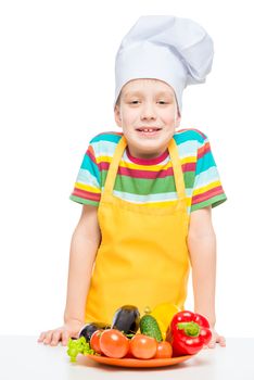 boy in a chef's cap and apron with a plate of vegetables isolated on white background
