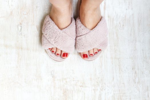 female legs with red nails in home fur fluffy pink slippers on a light wooden background. flat lay. Top view. The concept of a cozy bright girl house.