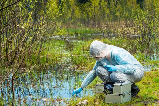 The ecologist takes a sample of water from a forest reservoir in protective clothing