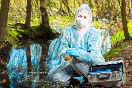 portrait of an ecologist in protective clothing while working, taking water samples from a forest river for analysis