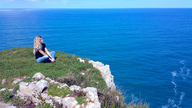 blonde girl sitting on the edge cliff, looking on the water