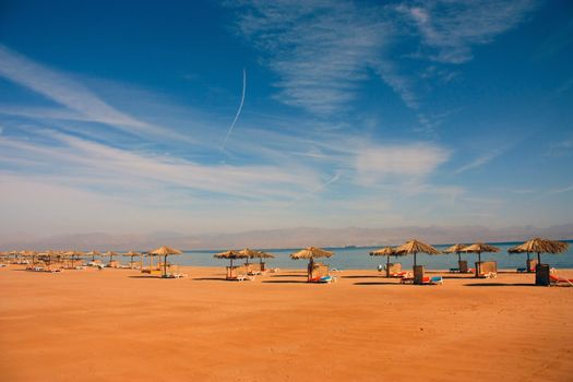 straw umbrellas from the sun on a beach on the shore of the red sea against a blue sky with white clouds and in the distance mountains in a haze on a sunny day.