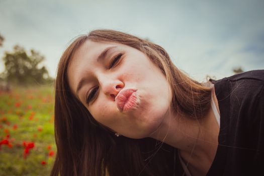 Young woman making duckface kiss while taking selfie picture with her smartphone or camera in field of red poppies. She having fun doing silly and funny to the camera.