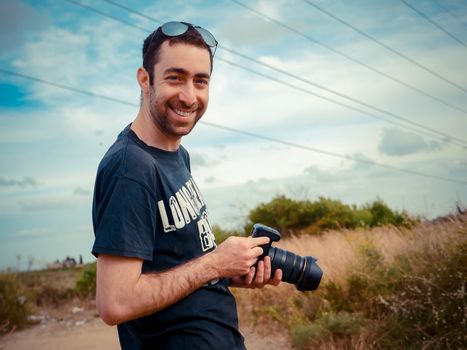 Happy young caucasian man photographer holding a digital camera in his hand and smiling to the camera outdoor in the field.