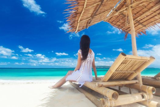 Woman sitting at chaise lounge with straw parasol on white sandy beach at Philippines