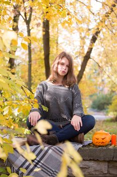 Beautiful teenager girl sitting in autumn garden on the fence , on woolen plaid blanket with halloween pumpkin and candles
