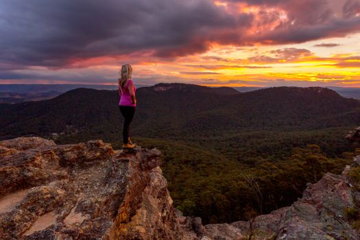 Storm clouds hover over Blue Mountains and valleys as the sun sets.  A woman stands on the rocky precipice taking in the magnificent views