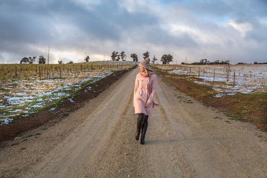 A woman walking down a country dirt road in winter.  She is wearing a thick woolen double breasted pale pink coat over jeans, a beanie and long boots.  There is light snow on the ground.