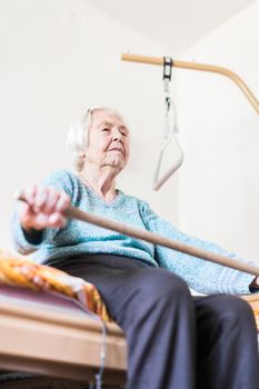 Elderly 96 years old woman exercising with a stick sitting on her bad. Geriatric health care home assisted support for older people concept. Care for the elderly.