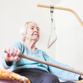 Elderly 96 years old woman exercising with a stick sitting on her bad. Geriatric health care home assisted support for older people concept. Care for the elderly.