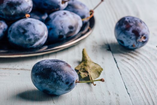 Ripe plums on metal plate and light wooden surface with leaves