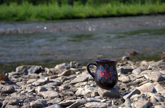 A black ceramic cup with a drink stands on the rocks on the bank of a mountain river. Altai, Siberia, Russia.