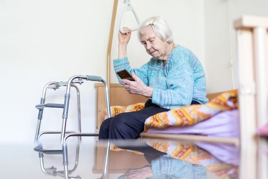 Elderly 96 years old woman reading phone message while sitting on medical bed supporting her by holder. Senior woman using wireless internet connection on smart phone.