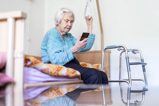 Elderly 96 years old woman reading phone message while sitting on medical bed supporting her by holder. Senior woman using wireless internet connection on smart phone.