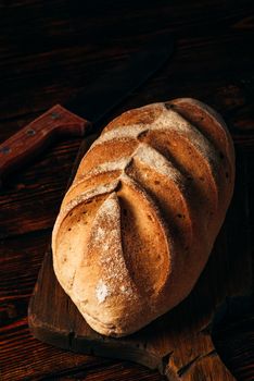 Homemade rye bread on cutting board with knife over dark wooden background.