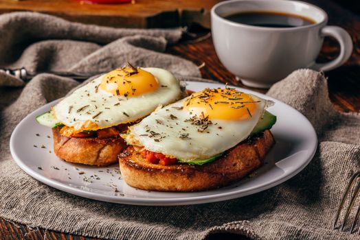 Italian toasts with vegetables and fried eggs on white plate and cup of coffee over grey rough cloth.