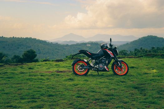 An orange sports motor bike resting on a green hill with beautiful mountains in the background