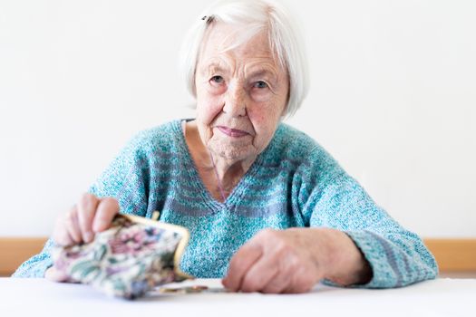 Concerned elderly 96 years old woman sitting at table at home and counting remaining coins from pension in her wallet after paying bills. Unsustainability of social transfers and pension system.