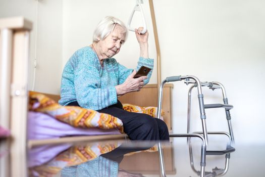 Elderly 96 years old woman reading phone message while sitting on medical bed supporting her by holder. Senior woman using wireless internet connection on smart phone.