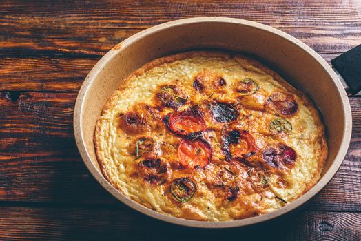 Cooked frittata with chorizo, tomatoes and chili peppers in a frying pan over dark wooden background. High angle view.