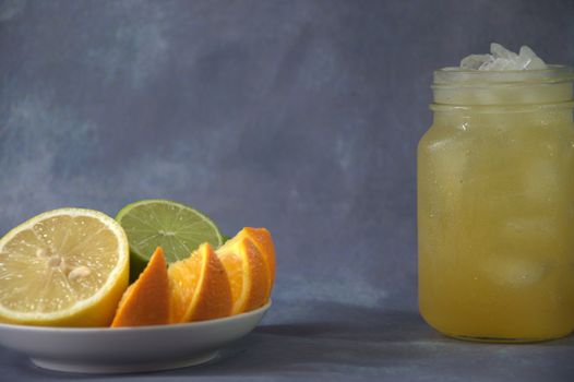 A citrus platter of lemon, orange and lime slices on a saucer and a glass mug with citrus juice and ice stand nearby.