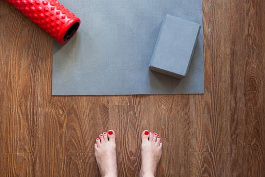 Woman is standing barefoot on floor in front of gymnastic mat and roller, she is going to do morning exercise complex. Only feet are visible. Healthy lifestyle, sport, weight loss concept. Horizontal.