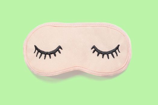 Pastel pink sleep mask with closed eyes embroidered on it with eyelashes on pastel green background. Top view, flat lay. Concept of vivid dreams. Accessories for girls and young women. Minimal style.