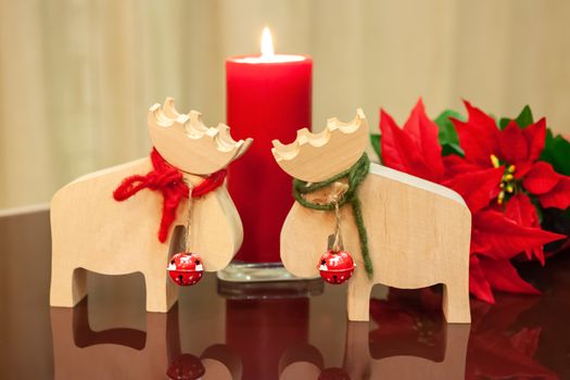 Christmas decor in modern interior. Scandinavian style, hygge. Christmas toys moose deer with red and green ropes tied around neck and bells with lit candle and with poinsettia in pot on glass table.