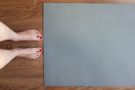 Woman is standing barefoot on floor in front of gymnastic mat, she is going to do morning exercise complex. Only legs are visible. Healthy lifestyle, weight loss concept. Horizontal. Copy space.