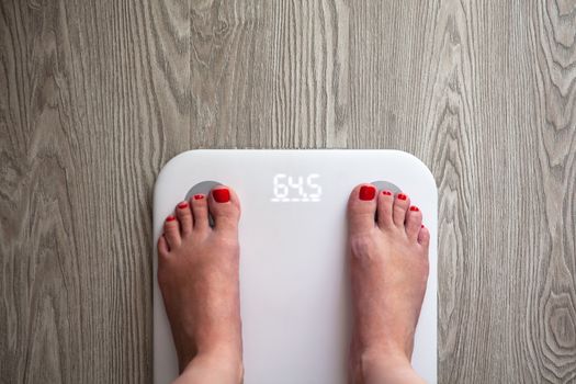 Woman stands on white modern electronic scales, which show 64.5 kg. Only feet are visible. Scales stand on gray wooden floor. Copy space. Healthy lifestyle, diet, weight loss concept. Top view.